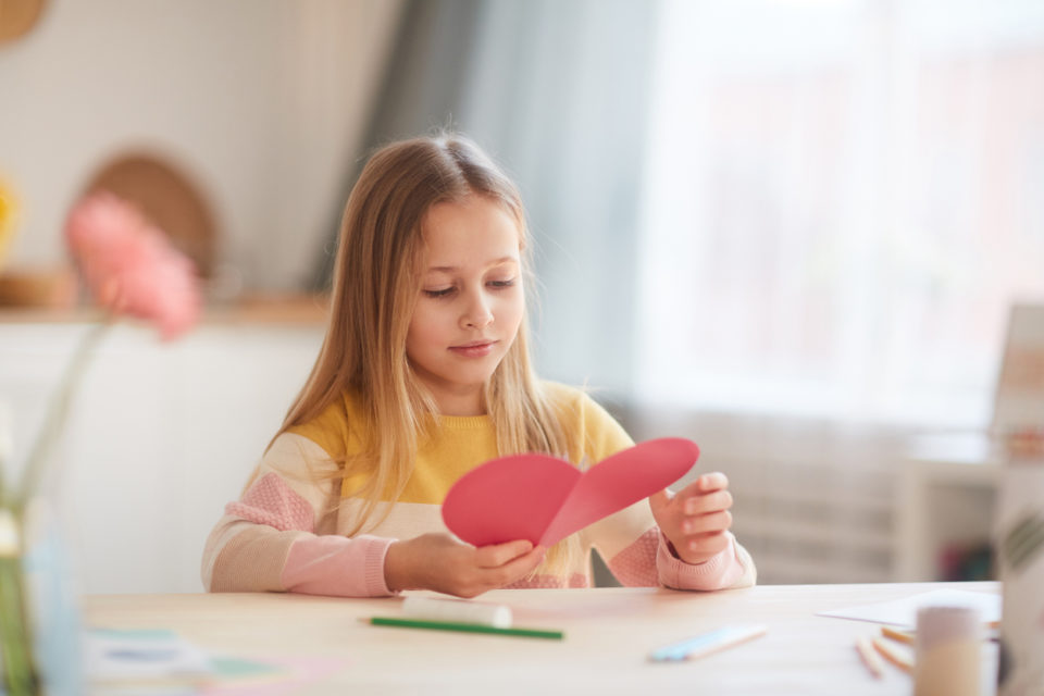 Portrait of girl holding heart-shaped card and smiling while sitting at table in cozy home interior