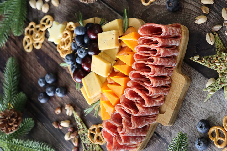 Image of Christmas tree shaped wooden charcuterie board covered with pretzels, blueberries, cherries, cheddar, apricots, Red Leicester cheese, salami slices, topped with cheese star, spruce needles, pistachios, pine cones, focus on foreground